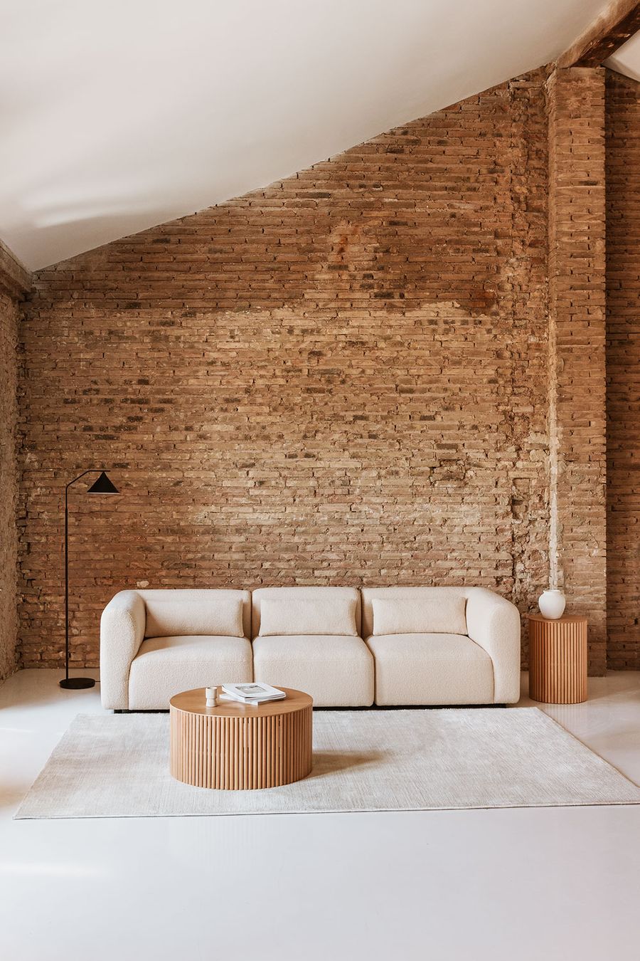 Canapé Fogler trois places beige devant un mur de briques, avec table basse en bois cannelé et lampadaire.