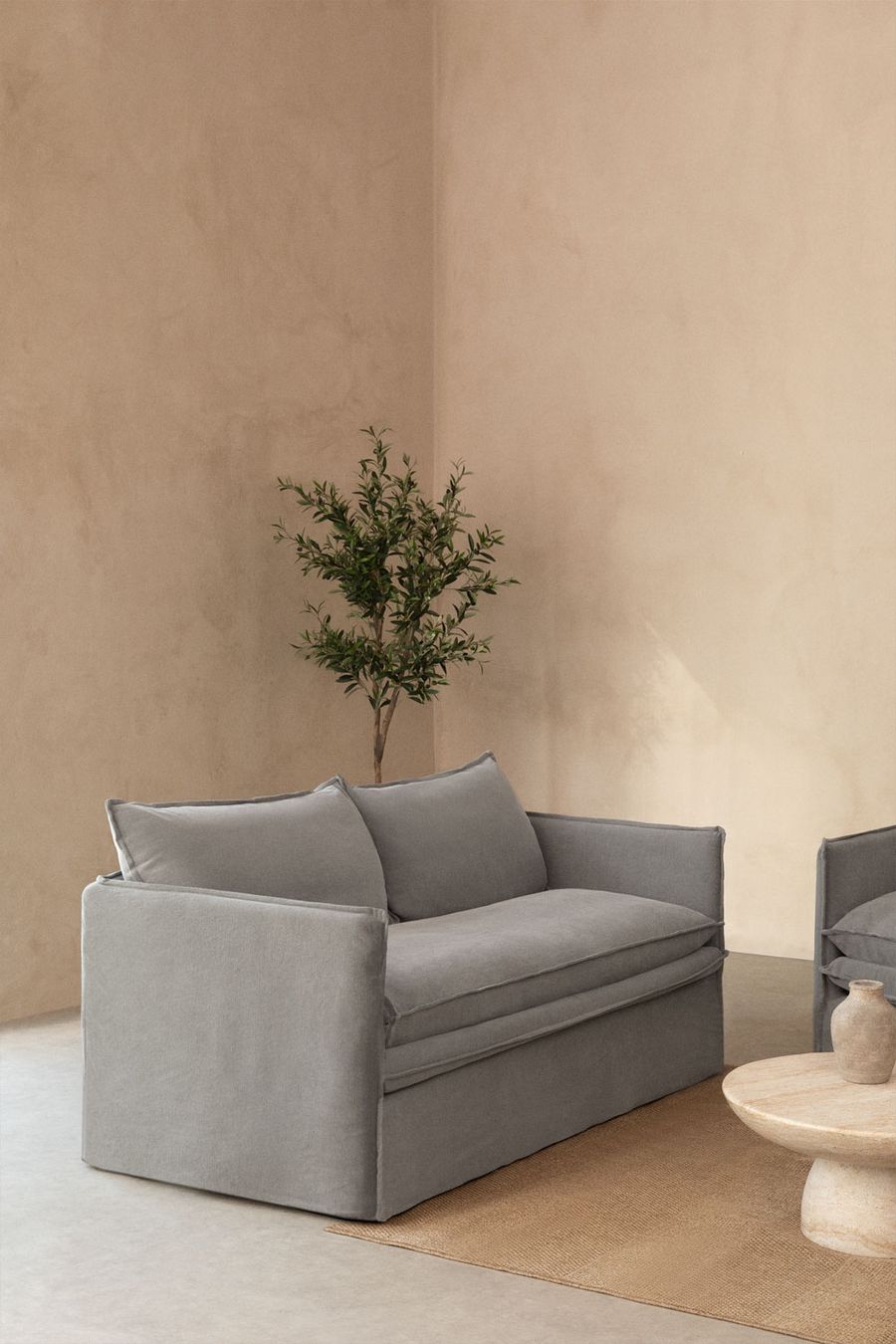 Canapé Grace gris dans un coin de pièce aux murs beige, avec un olivier et une table basse.