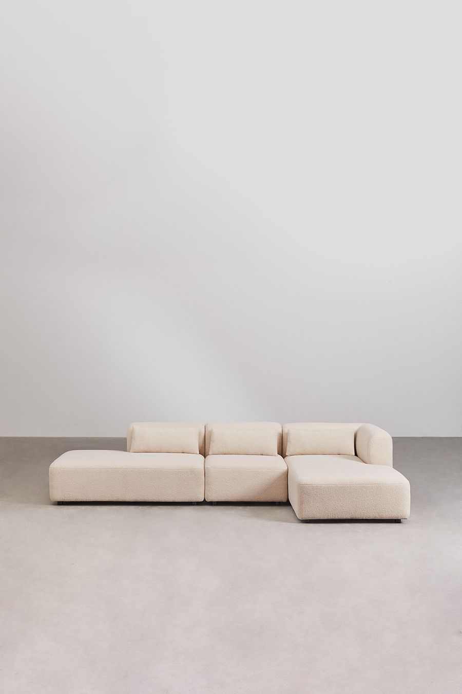 Canapé Fogler d'angle beige bouclé, méridienne à gauche, sur sol béton contre un mur blanc nu.