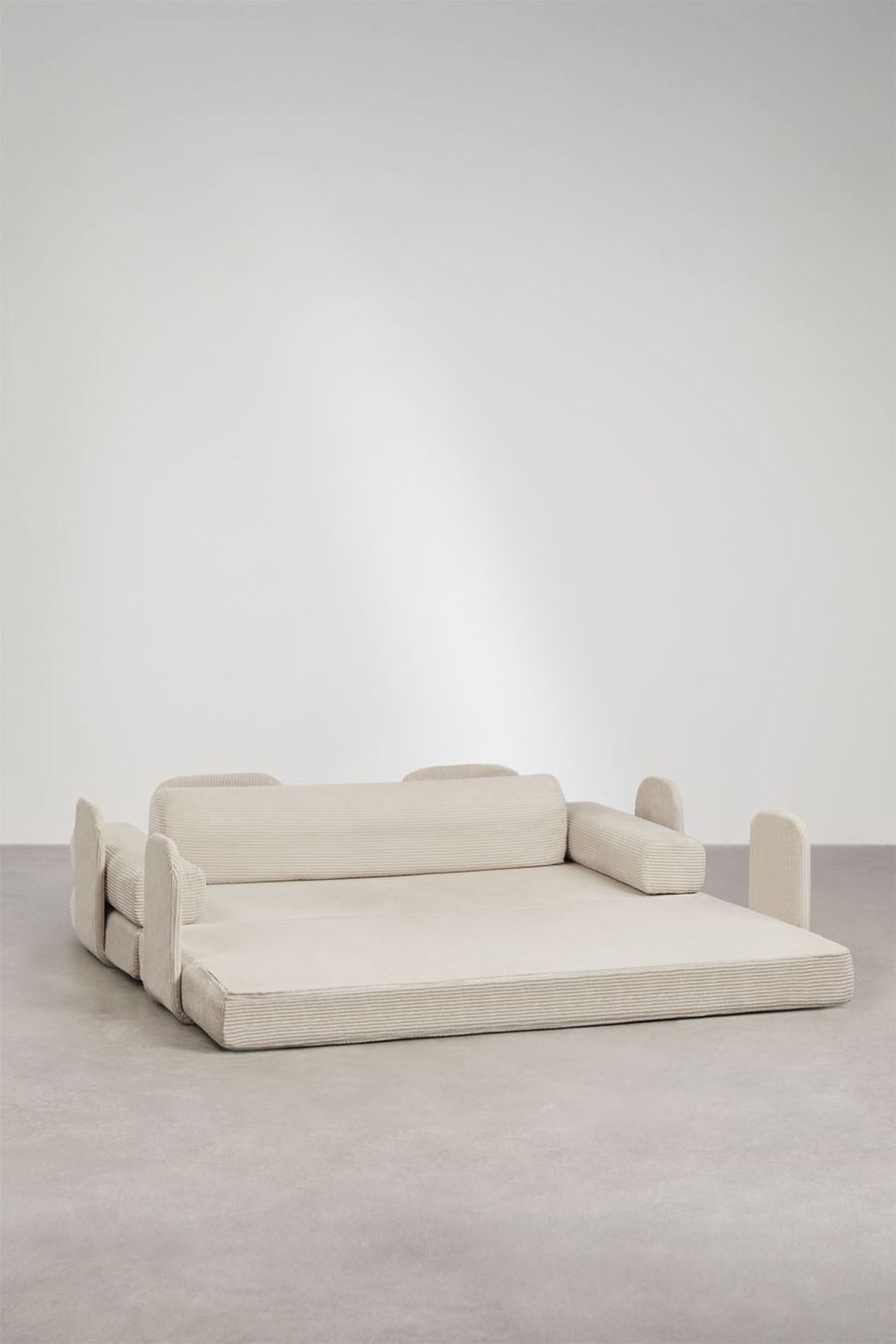 Canapé Paoly déplié en lit, modules d'accoudoirs posés à part autour de l'assise beige sur sol gris.
